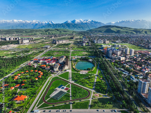 Wide aerial view of a large green park area with a running track and lake in Bishkek, backed by snow-capped Tian Shan mountains