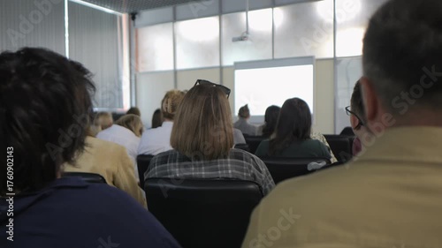 Group of people are sitting in a classroom. A man is sitting in the front row. A woman is sitting in the back row