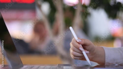 Person is holding a white pen in their hand. The pen is on a laptop. The laptop is on a table
