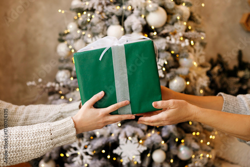 Two hands exchanging a beautifully wrapped green gift box with a silver ribbon in front of a decorated Christmas tree, capturing the spirit of giving and holiday joy