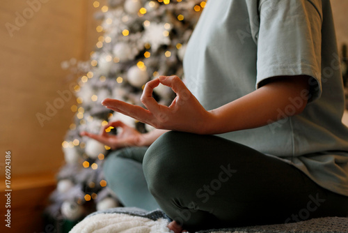 Individual practicing meditation in a serene indoor setting, seated cross-legged on a soft surface, with a beautifully decorated Christmas tree in the background, promoting relaxation and mindfulness