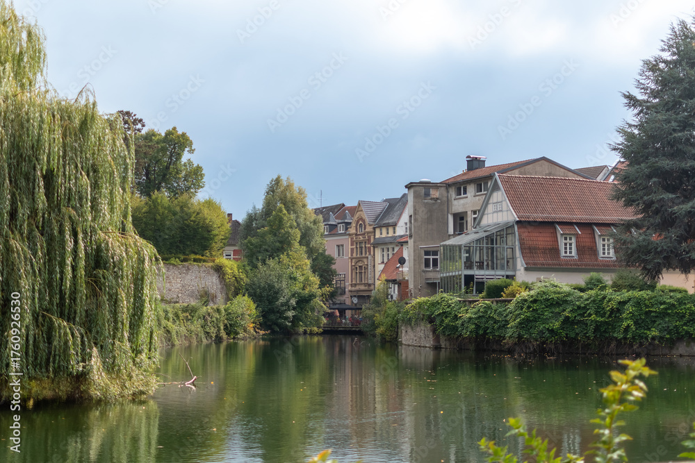 Obraz premium Tranquil canal or river with reflections, lined by lush green trees, an old stone wall, and a row of traditional European houses under a slightly overcast sky