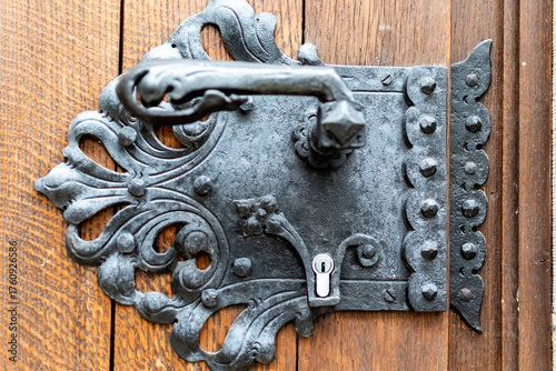 Close-up image of an elaborate, antique black wrought iron door handle and lock plate, mounted on a rustic wooden door with visible grain