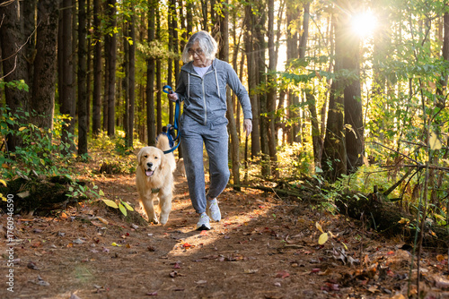 Senior woman walking golden retriever through pine forest in autumn sunlight