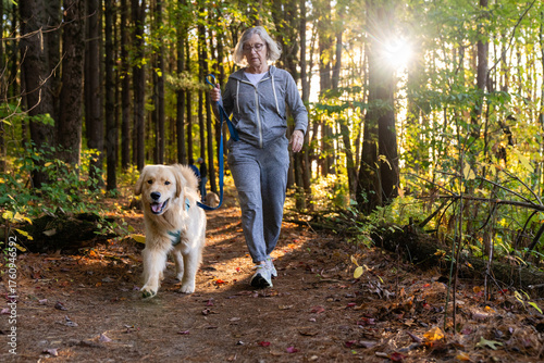 Active senior woman walking golden retriever on forest trail in autumn sunlight