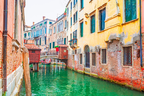 Fototapeta Naklejka Na Ścianę i Meble -  Narrow canal in Venice, Italy, with historic buildings on either side. A small arched bridge is visible in the distance, and the buildings are reflected in the green water