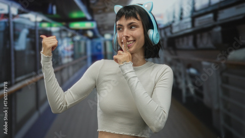 Young hispanic woman with cat ear headphones makes finger to lips for silence in a building corridor; quiet. Free Download Young hispanic woman with cat ear headphones makes finger to lips for silence in a building corridor; quiet. - GFXLoad