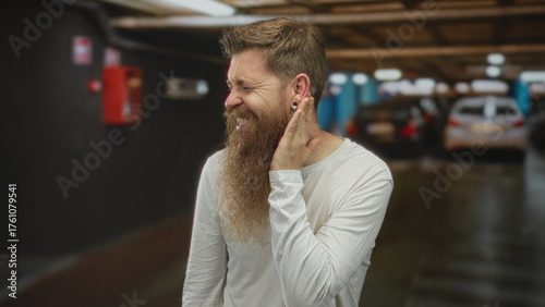 Man with bushy beard and casual white shirt holds hand to ear in building filled with blurred cars reflecting overhead lights; discomfort.