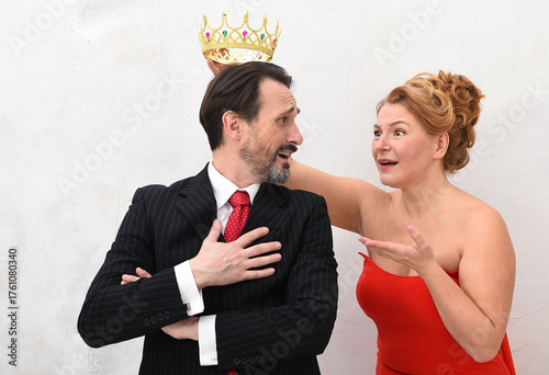 A woman playfully places a crown on a man's head while he looks up at her with a surprised and amused expression