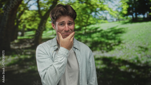 Young hispanic man with hand on cheek for toothache in green park under trees and sunlight; pain regret.