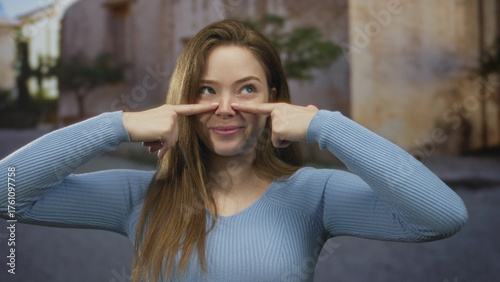 Woman with long hair and blue sweater smiling points finger to nose on sunlit old town cobblestone street; playful moment.