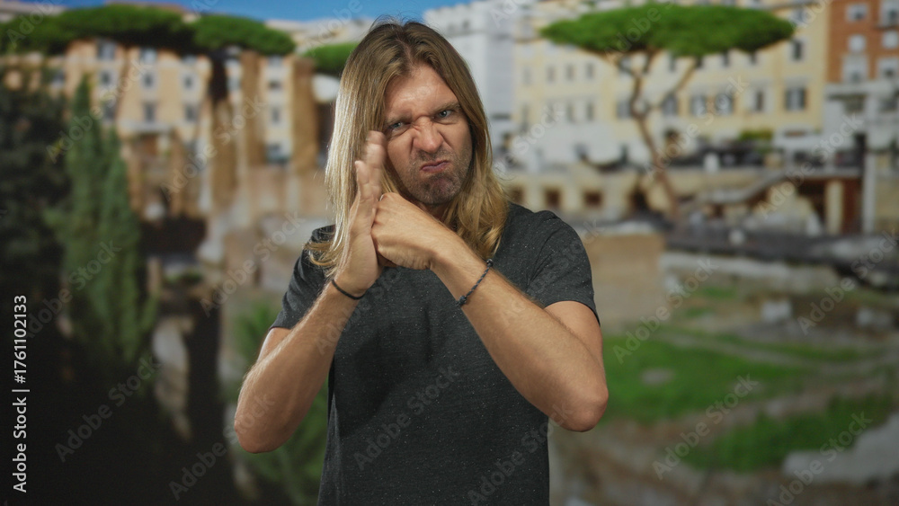 Fototapeta premium Blond man holds fist under palm in blocking gesture inside a building with ancient roman ruins in the background; defiance.