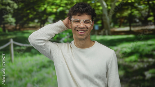 Man scratching head and smiling while standing in a green forest setting casually wearing glasses; serenity.