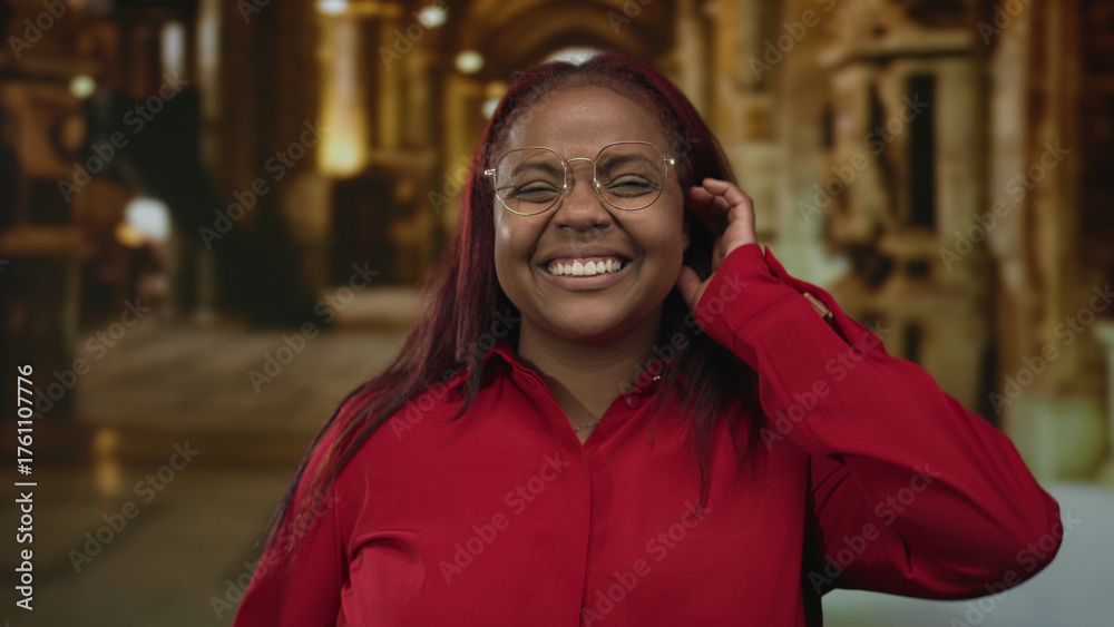 Fototapeta premium Woman in red shirt and round glasses smiles and touches ear amid stained glass columns in dimly lit church building; warmth joy.