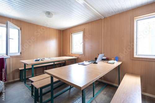 A workers' canteen inside a modular cabin, with long tables and benches for meals. A laptop suggests the space is also used for breaks or work during downtime at a construction site.