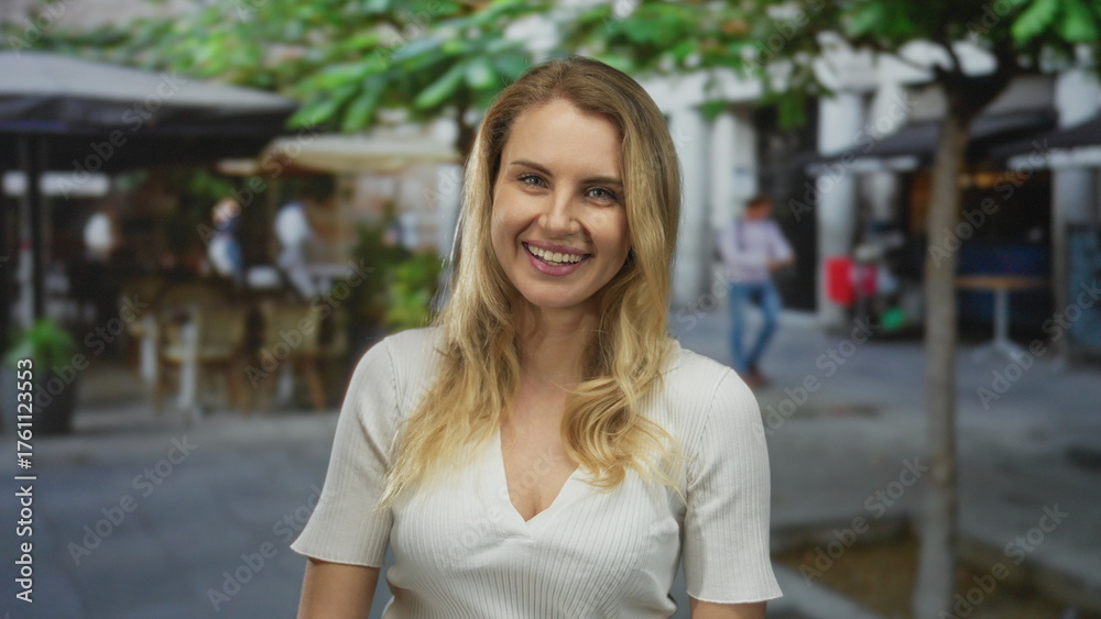 Fototapeta premium Woman smiling on urban street with blonde hair wearing white shirt outdoors in city setting showing casual happiness in lively environment under sunny sky with blurred background.
