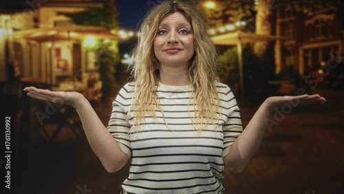 Wallpaper Mural Young blonde woman shrugging with palms up on a dimly lit cobblestone street at night; uncertainty and curiosity. Torontodigital.ca