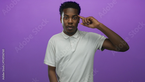Photography Young african american man in white polo shirt points finger to temple in studio against purple studio wall; playful madness