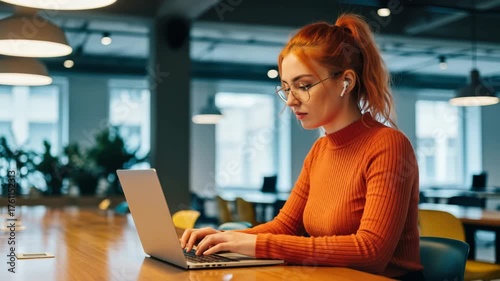 A young woman with vibrant red hair in a ponytail, wearing glasses and an orange turtleneck, focuses on her laptop while wearing wireless earbuds in a contemporary office setting