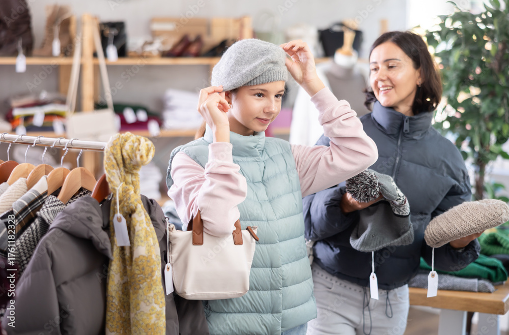 Obraz premium Teen girl is fitting on and trying on winter hat in store, family shopping with mom. Buyer looks at herself in mirror while shopping, evaluates her appearance. Fitting clothes and accessories.