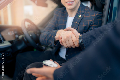 Young male in suit finalizing car purchase with handshake in dealership, sunlight