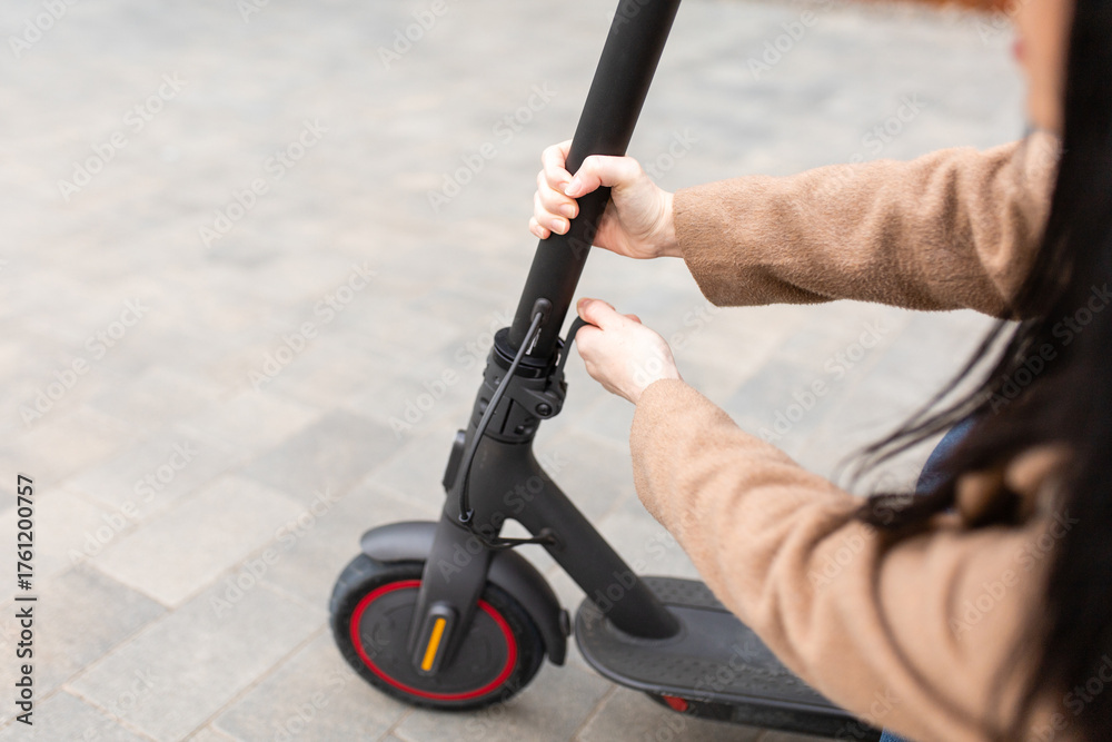 Obraz premium Young woman in a beige coat folds an electric scooter in the middle of a city street. A person uses an environmentally friendly form of transport in the form of an electric scooter.