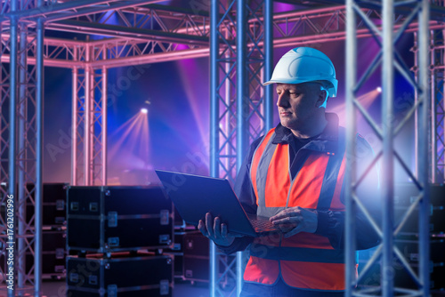 Stage construction supervisor with laptop monitors truss installation, managing lighting structures, safety, and technical setup for live events.
