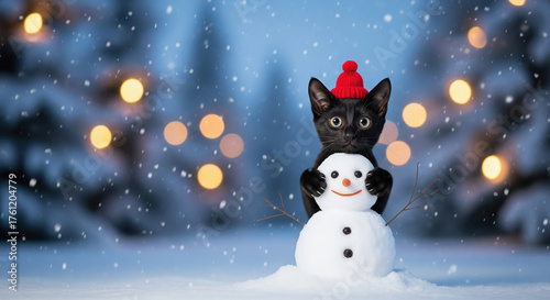 Cute black cat in a red hat building a tiny snowman in a winter snow scene.