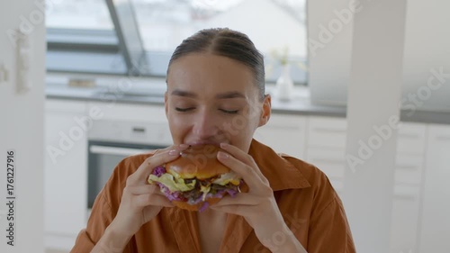 A young woman sits at a bright kitchen table, savoring a large burger filled with fresh ingredients.