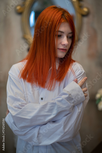 A beautiful girl with long red hair in a white formal classic shirt or blouse hugs herself by the shoulders. Shirt or blouse is badly wrinkled
