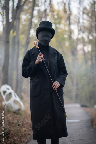 A man with a face in black bandages is dressed in an old vintage style: a classic black coat and tweed vest, a top hat, a walking stick with a skull in his hands. The invisible man or the zombie
