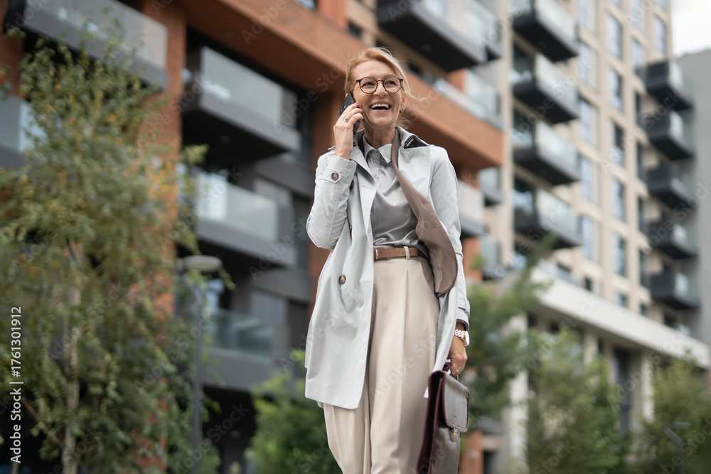 Fototapeta premium View of happy, smiling, business female . Confident senior woman in business outfit talking on smartphone near office center. Outdoors. Daily life. Routine. Urban design