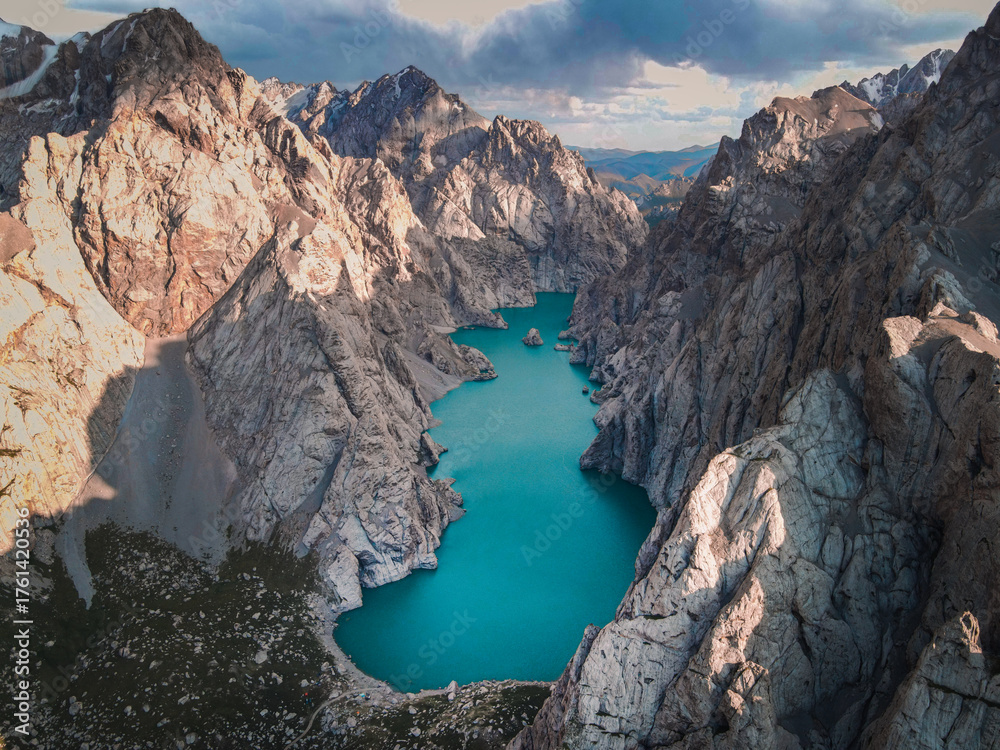 Fototapeta premium High-angle aerial view of the vibrant turquoise Kol Suu Lake nestled deep in a massive, rocky canyon under a dramatic, colorful sky