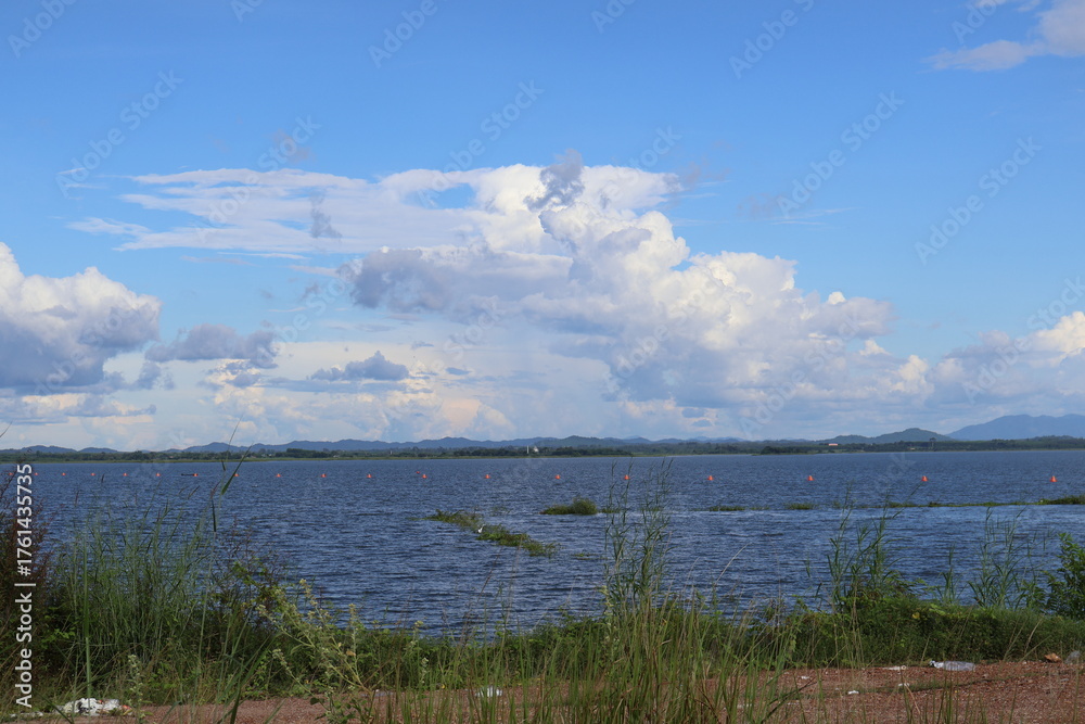 Fototapeta premium A serene lakeside view under a partly cloudy sky. In the foreground, there's an expanse of calm water reflecting the clouds above