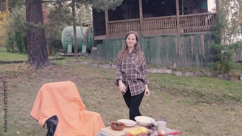 A young woman wearing a brown plaid shirt enjoys a delightful autumn picnic outside the city. Surrounded by nature, she expresses her happiness while standing by a table laid out with food.