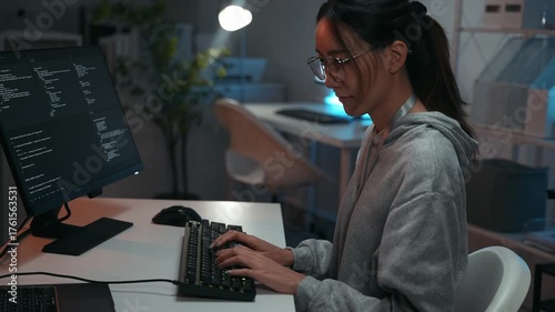 Focused Asian woman programmer working late at desk, typing on a keyboard with code on the computer screen. Developing complex software for data and cyber security in a dark office