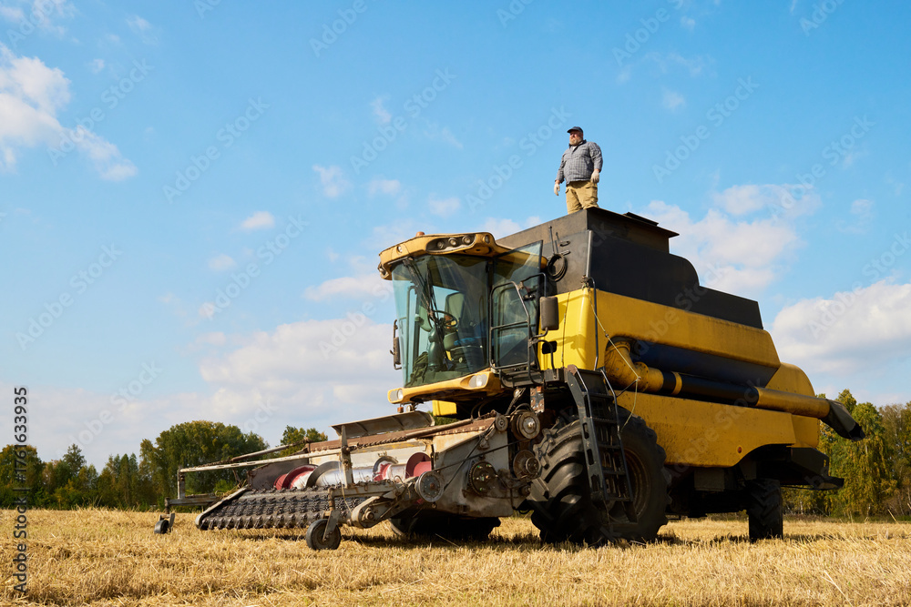 Obraz premium Caucasian middle aged man standing on top of large agricultural combine harvester in open field during harvest season, observing farmland and overseeing machinery operation