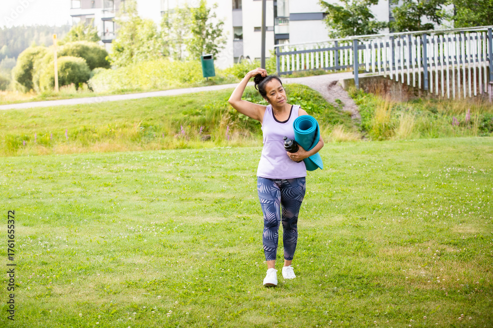 Obraz premium Middle aged woman of color walking with yoga mat to park, embracing diversity, wellness, and active modern lifestyle in nature.