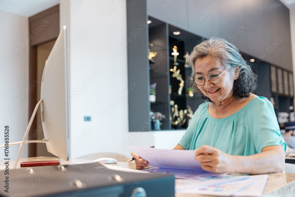 Obraz premium Senior woman working from home using a desktop computer, analyzing documents and taking notes. Concept of active retirement, lifelong learning, remote work, and digital literacy among seniors.
