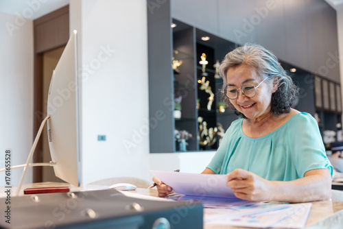 Senior woman working from home using a desktop computer, analyzing documents and taking notes. Concept of active retirement, lifelong learning, remote work, and digital literacy among seniors.