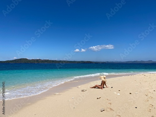 A woman sitting on a beautiful tropical beach in Madagascar with clear turquoise water and white sand. The bright sunlight, blue sky, and crystal-clear ocean create a peaceful and exotic summer atmos