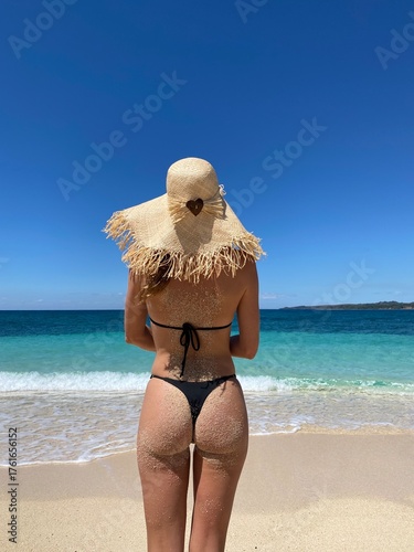 A woman standing on a beautiful tropical beach in Madagascar with clear turquoise water and white sand. The bright sunlight, blue sky, and crystal-clear ocean create a peaceful and exotic summer 