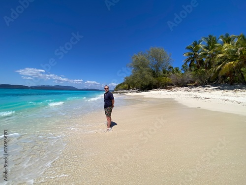 A man standing on a tropical island in Madagascar across clear turquoise waters. The bright sunlight reflects on the calm ocean, highlighting the white sandy shores and creating a pe