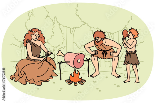 Family of primitive people stands around fire in forest, roasting meat for dinner or sewing up clothes. Group of primitive people of different ages living in stone age dressed in rags