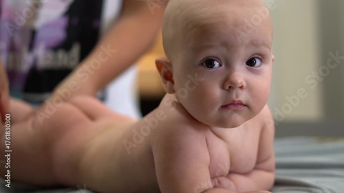 A baby is laying on a table with a woman touching it. The baby is looking at the camera with a surprised expression