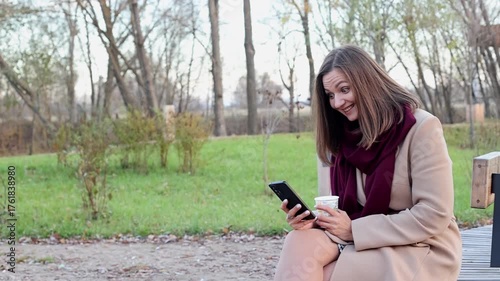 Female in beige coat and maroon scarf enjoys coffee while focused on smartphone, captured with a smooth dolly movement, showcasing serene outdoor environment