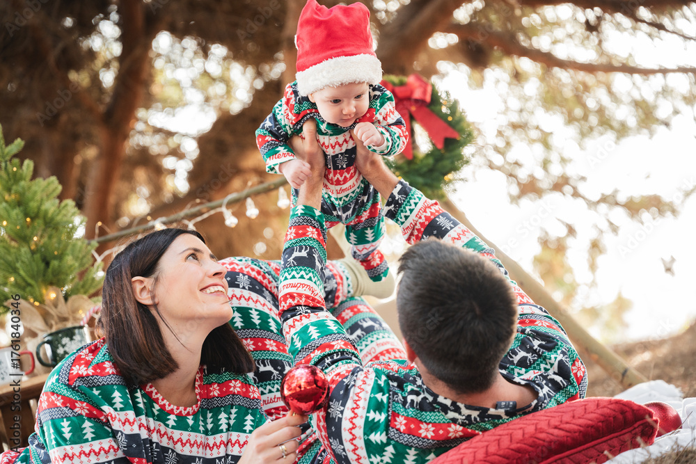 Obraz premium Parents lifting their baby in a santa hat, all wearing festive pajamas, creating joyful holiday memories outside