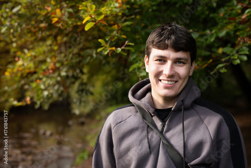 A young man is resting in an autumn park, smiling sincerely, fresh air, nature, walking and light jogging relieve his depression. A young guy wearing a grey hoodie stands near river