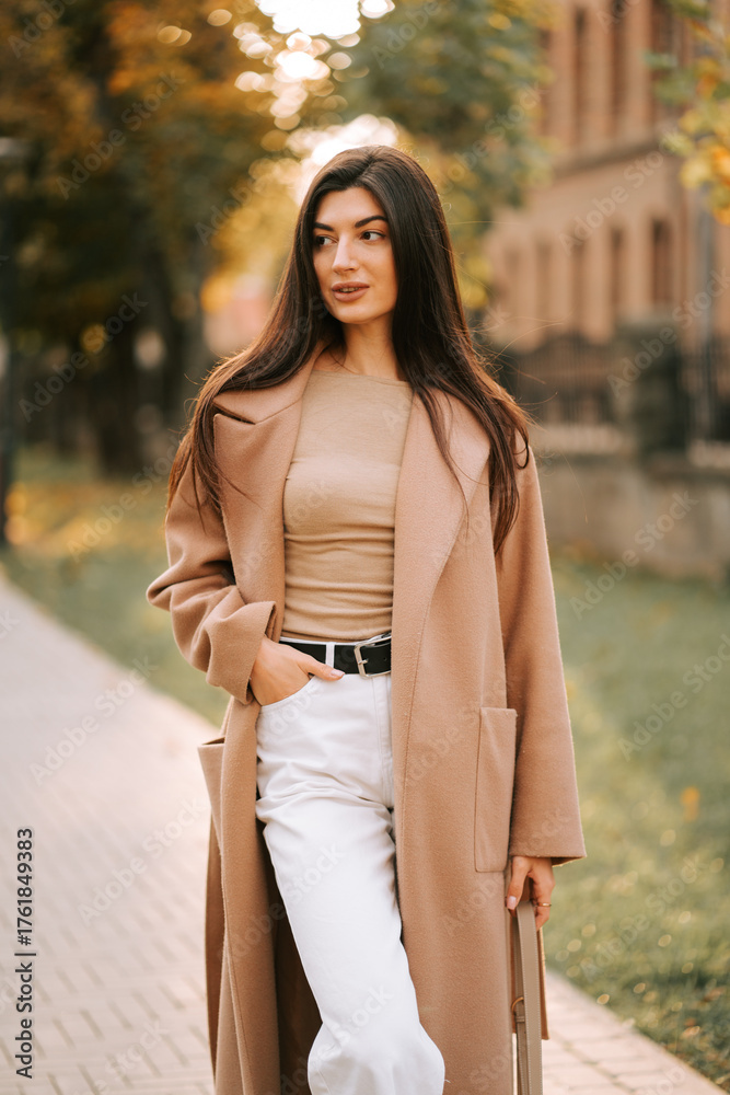 Fototapeta premium Stylish woman walking in autumn park wearing a long coat and white pants