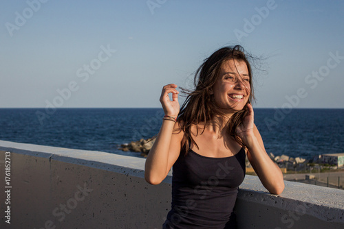 Cheerful suntanned young woman with sea and blue sky in background. Portrait of beautiful brunette girl laughing during seashore walk before sunset. Having good time on summer vacation concept.